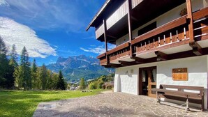 A building with a stone-paved courtyard, wooden balcony, and a bench. - Charming ground floor flat with mountain views (Cortina d'Ampezzo)