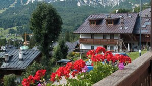 Vista sulle montagne con ringhiera in legno, fiori rossi e viola e un edificio con tetto spiovente.