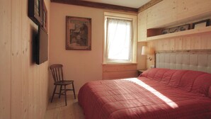 Un dormitorio con cabecera de madera, una cama individual con una colcha roja, una silla de madera, una ventana con cortinas blancas y un televisor fijado a la pared.