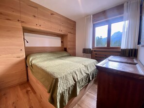 Un lit superposé en bois avec une couverture verte, un bureau en bois et une fenêtre donnant sur la montagne.