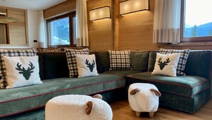 Un salon chaleureux avec un canapé d’angle vert agrémenté de coussins à carreaux et sur le thème des cerfs, deux poufs blancs et moelleux en forme de mouton, et un plancher en bois.