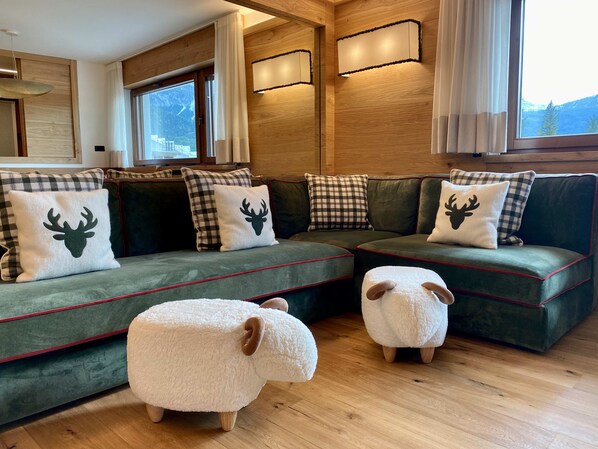 Un salon chaleureux avec un canapé d’angle vert agrémenté de coussins à carreaux et sur le thème des cerfs, deux poufs blancs et moelleux en forme de mouton, et un plancher en bois.