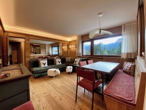 Une pièce chaleureuse avec un plancher en bois, un canapé agrémenté de coussins à motifs, une table à manger avec des chaises, et une grande fenêtre offrant une vue sur les montagnes.