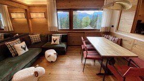 L’intérieur d’un chalet accueillant, avec un plancher en bois, un canapé vert agrémenté de coussins à carreaux, un pouf blanc en forme de mouton, une table à manger avec des chaises rouges et une grande fenêtre offrant une vue sur les montagnes.