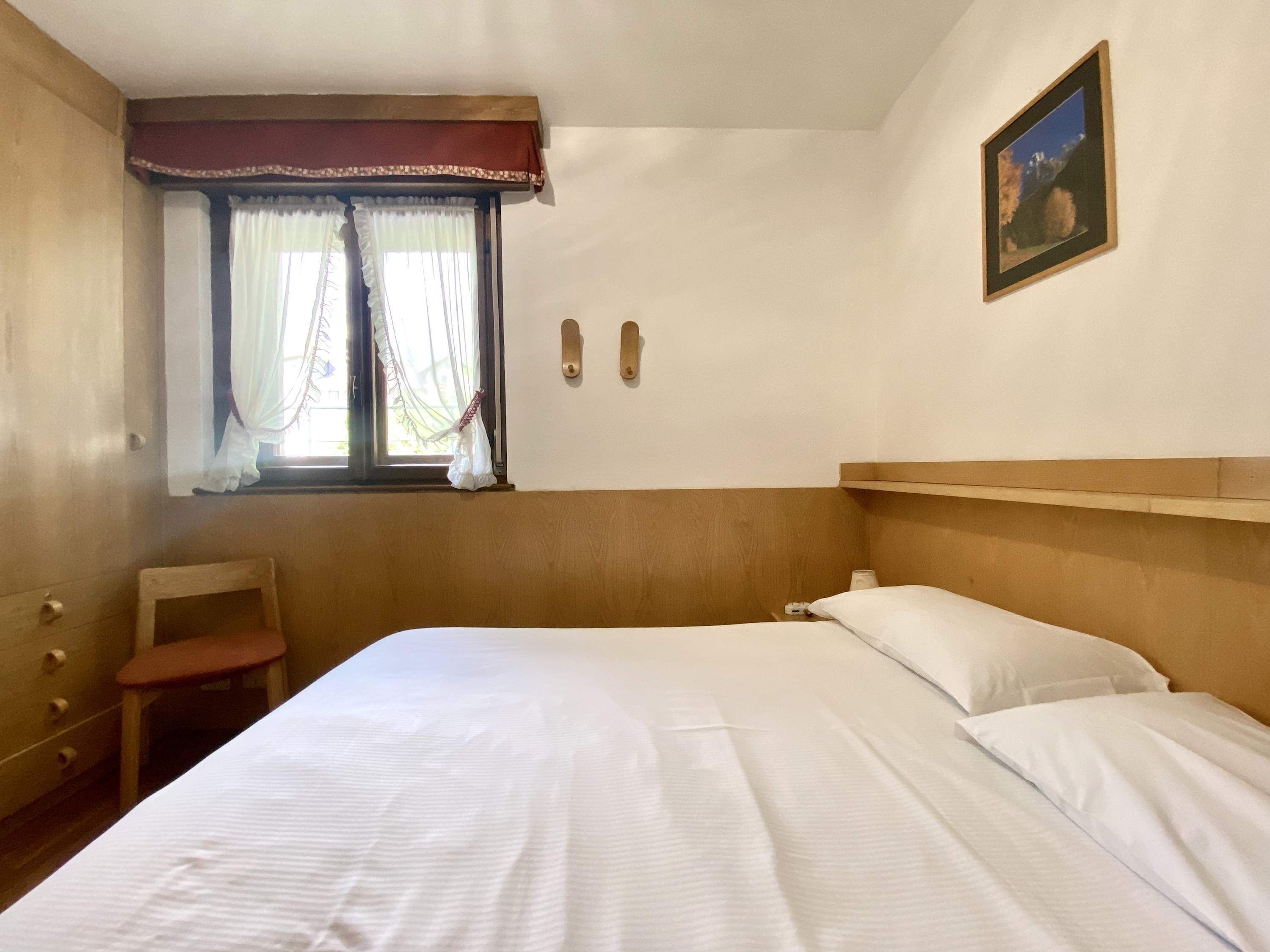 Ett hotellrum med en säng, en trägavel, en liten trästol, ett fönster med gardiner, en tavla som hänger på väggen och en hylla ovanför sängen.
