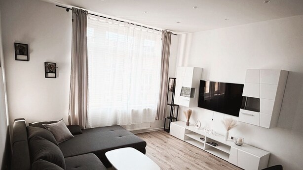 A modern living room with a sectional sofa, a coffee table, a TV mounted on a white cabinet, and a large window with curtains. - Vacation apartment - beautiful home Li-Apartments (Pforzheim)