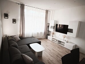 Un salon moderne avec un canapé d’angle, une table basse, un téléviseur fixé sur un meuble blanc et une grande fenêtre avec des rideaux.