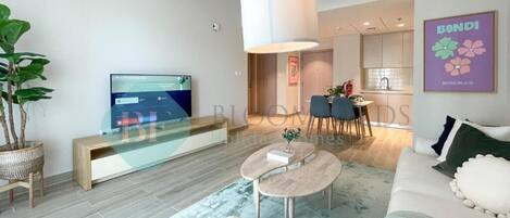 A modern living room with a sofa, a coffee table, and a TV. There is a dining area with chairs and a table in the background.