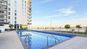 Una piscina con bordes de azulejos, una escalera metálica y un edificio blanco con balcones al fondo.