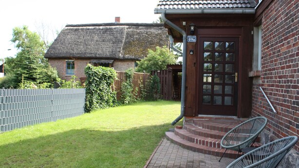 Une maison en briques avec un toit de chaume, une porte en bois et un petit porche avec deux chaises.