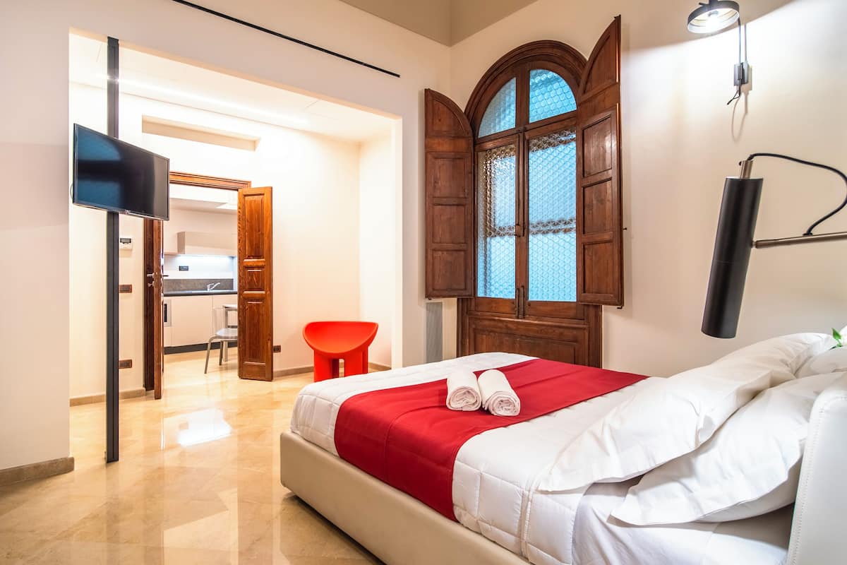 A hotel room with a bed, a red and white bedspread, a wooden arched window, a red chair, and a TV mounted on the wall.