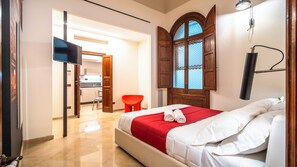 A hotel room with a bed, a red and white bedspread, a wooden arched window, a red chair, and a TV mounted on the wall.