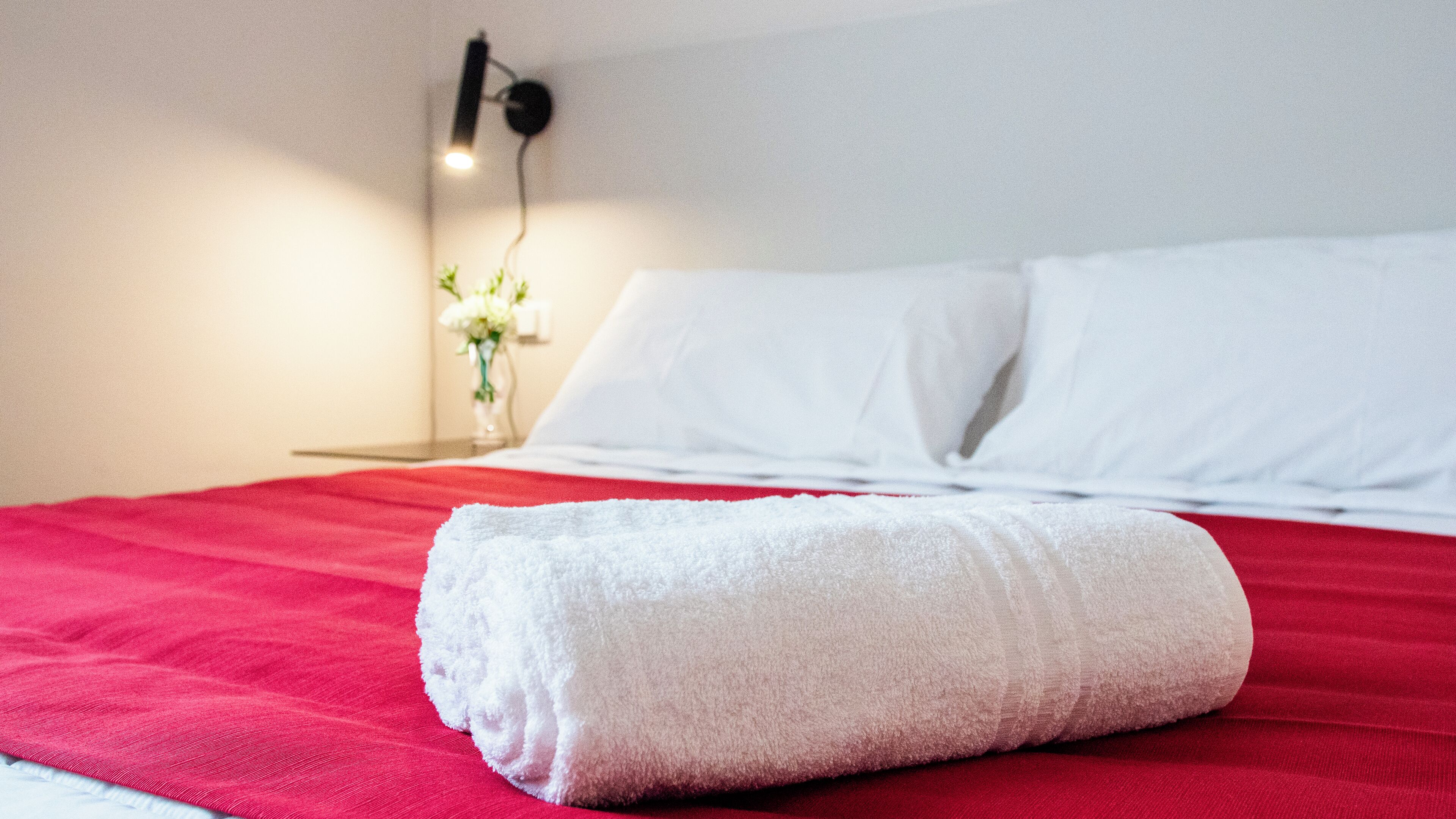 A neatly made bed with a red blanket and a rolled white towel on top.