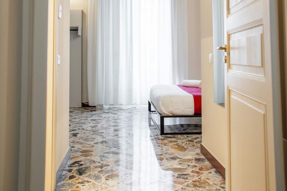 A narrow hallway with a bed at the end, a radiator, and a window with curtains.