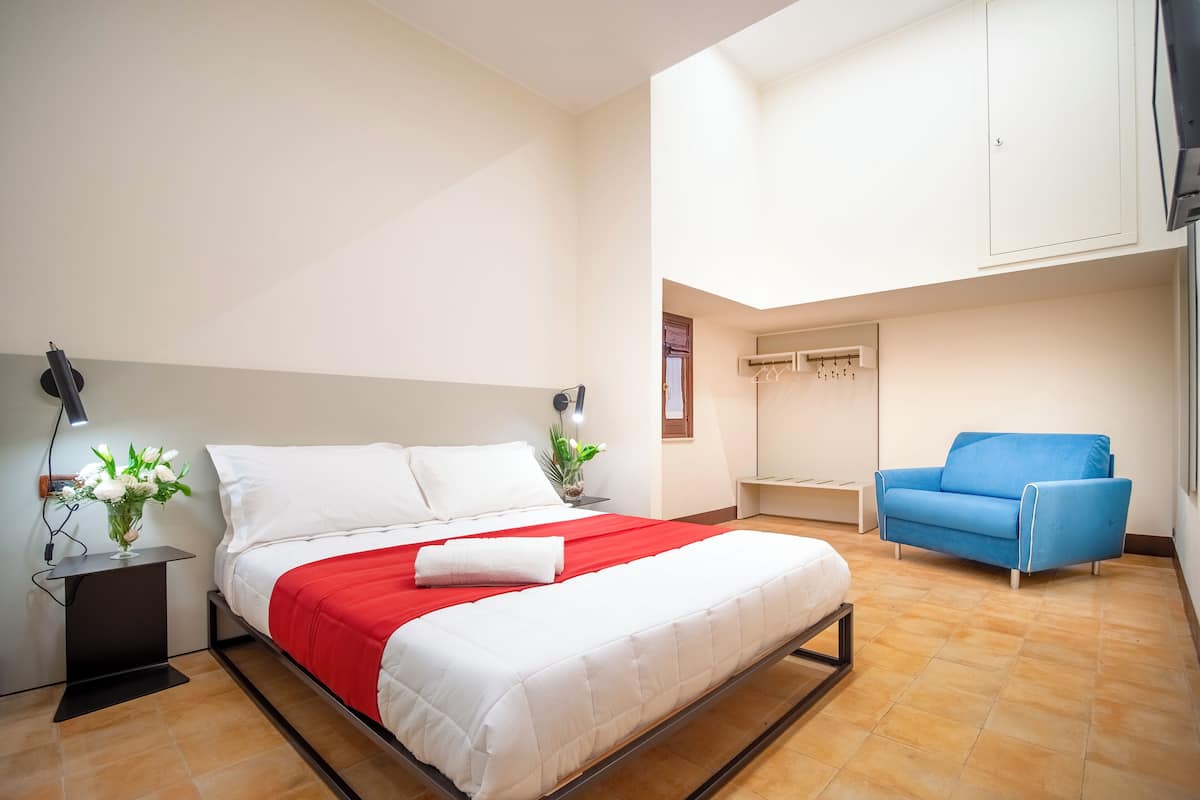 A modern hotel room with a bed, bedside tables, a desk, a blue sofa, and a wardrobe.