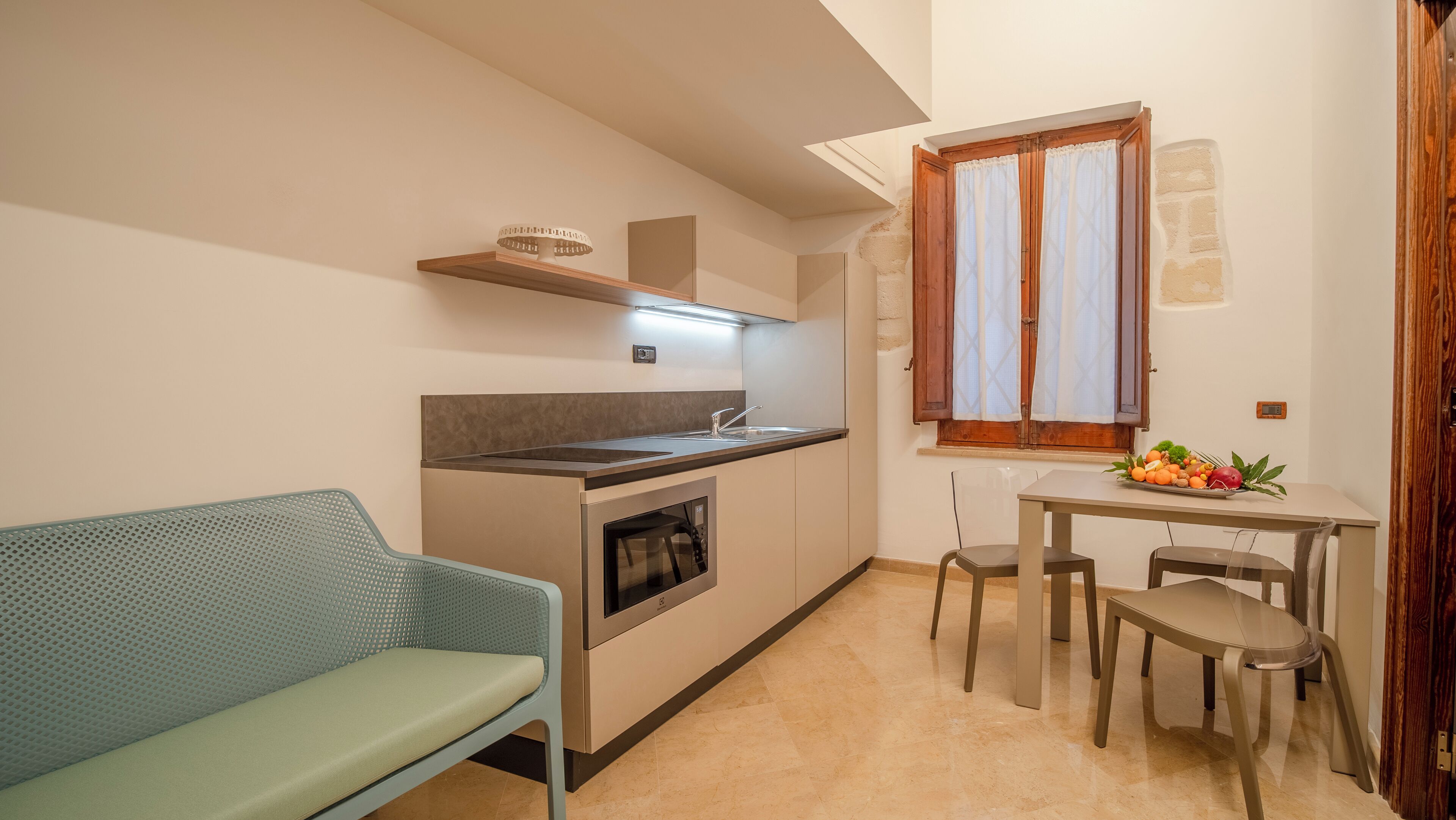 A compact kitchen with a built-in oven, a sink, and a window with curtains. There is a dining table with chairs and a small sofa in the corner.
