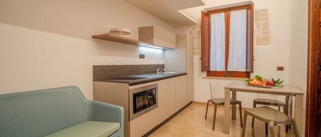 A compact kitchen with a built-in oven, a sink, and a window with curtains. There is a dining table with chairs and a small sofa in the corner.