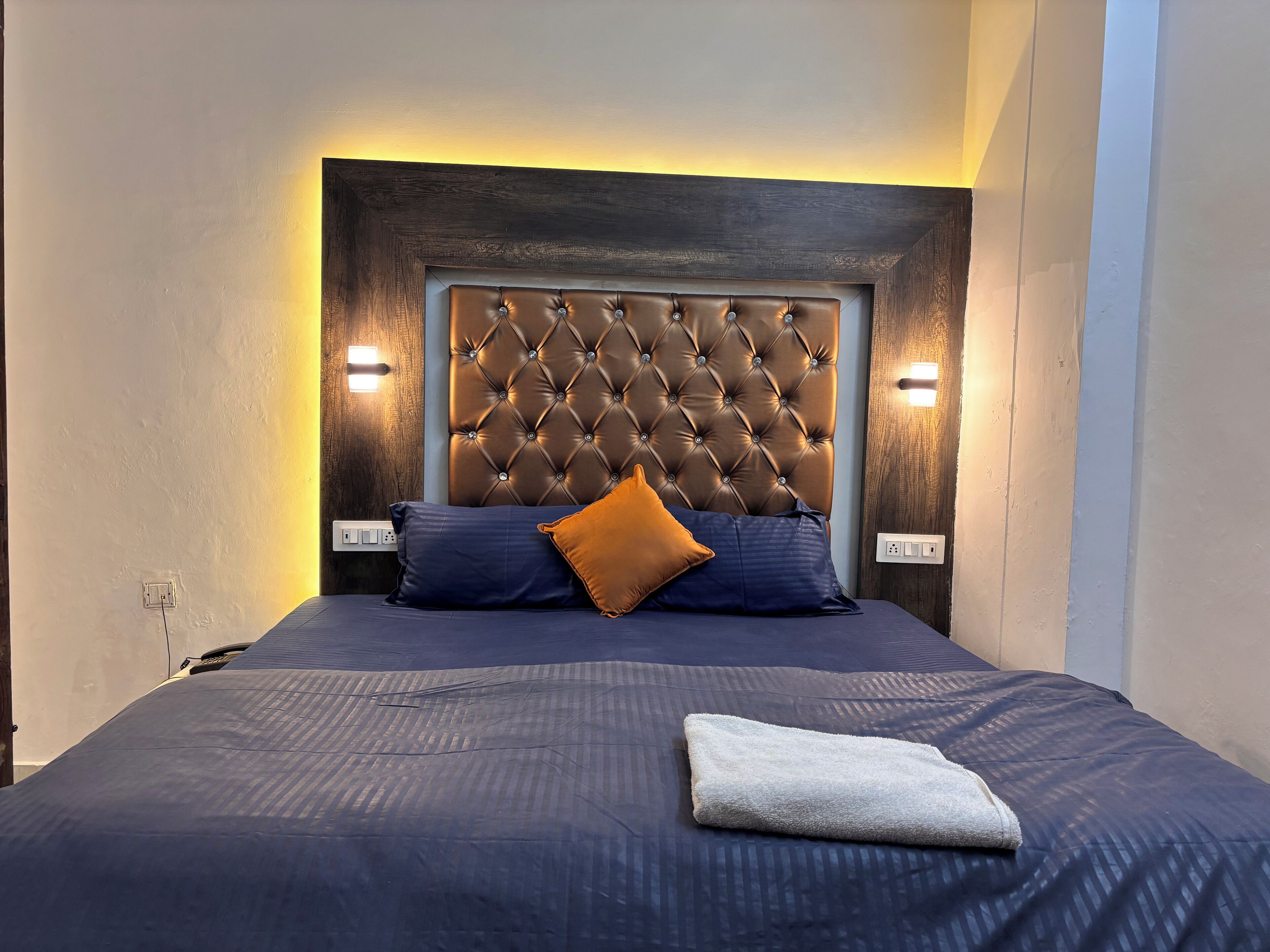 Un lit bien fait, avec une tête de lit capitonnée, deux lampes de chevet et une serviette blanche pliée posée sur le lit.