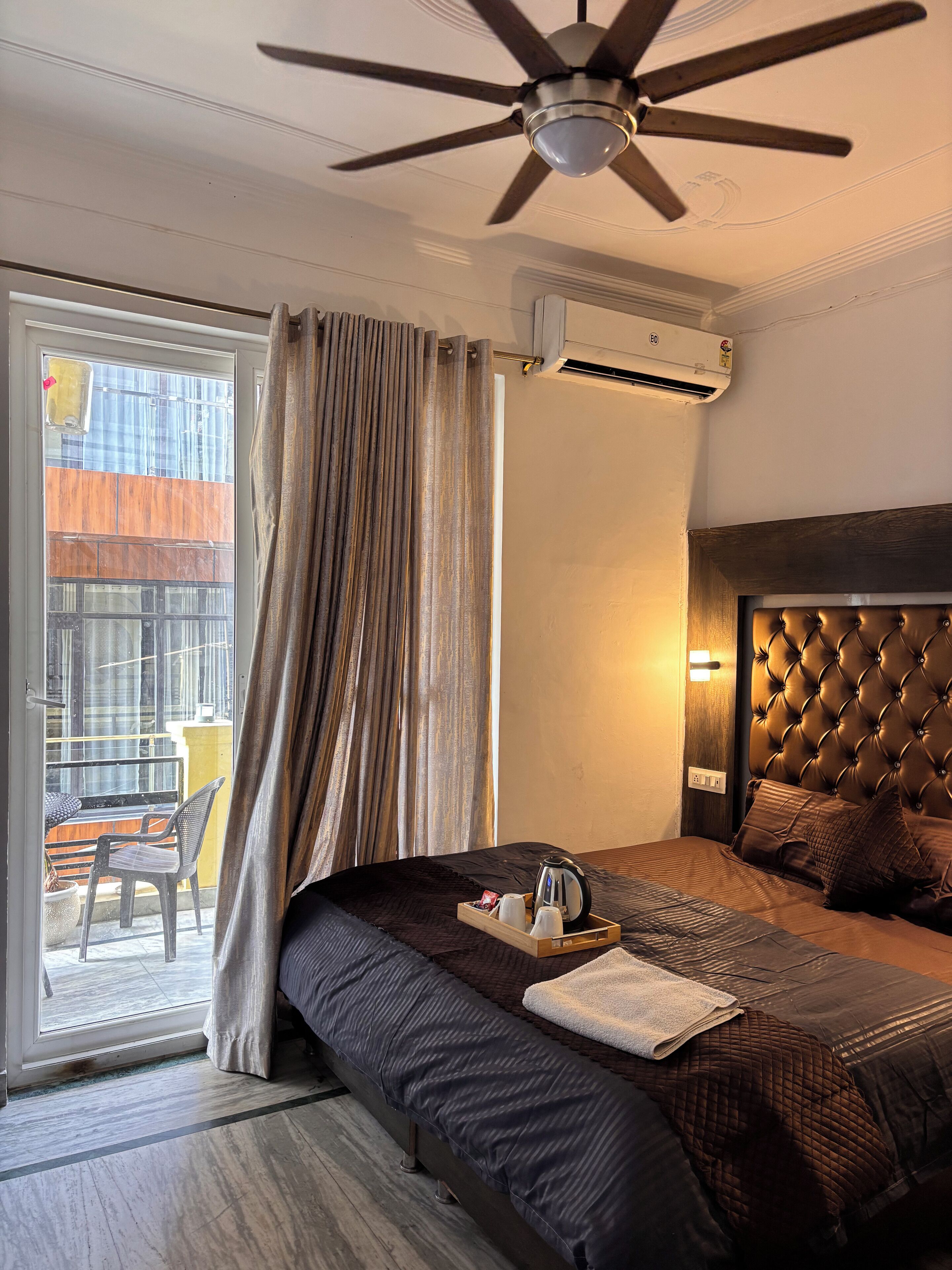 A hotel room with a bed, a ceiling fan, an air conditioning unit, and a view of the outside balcony.
