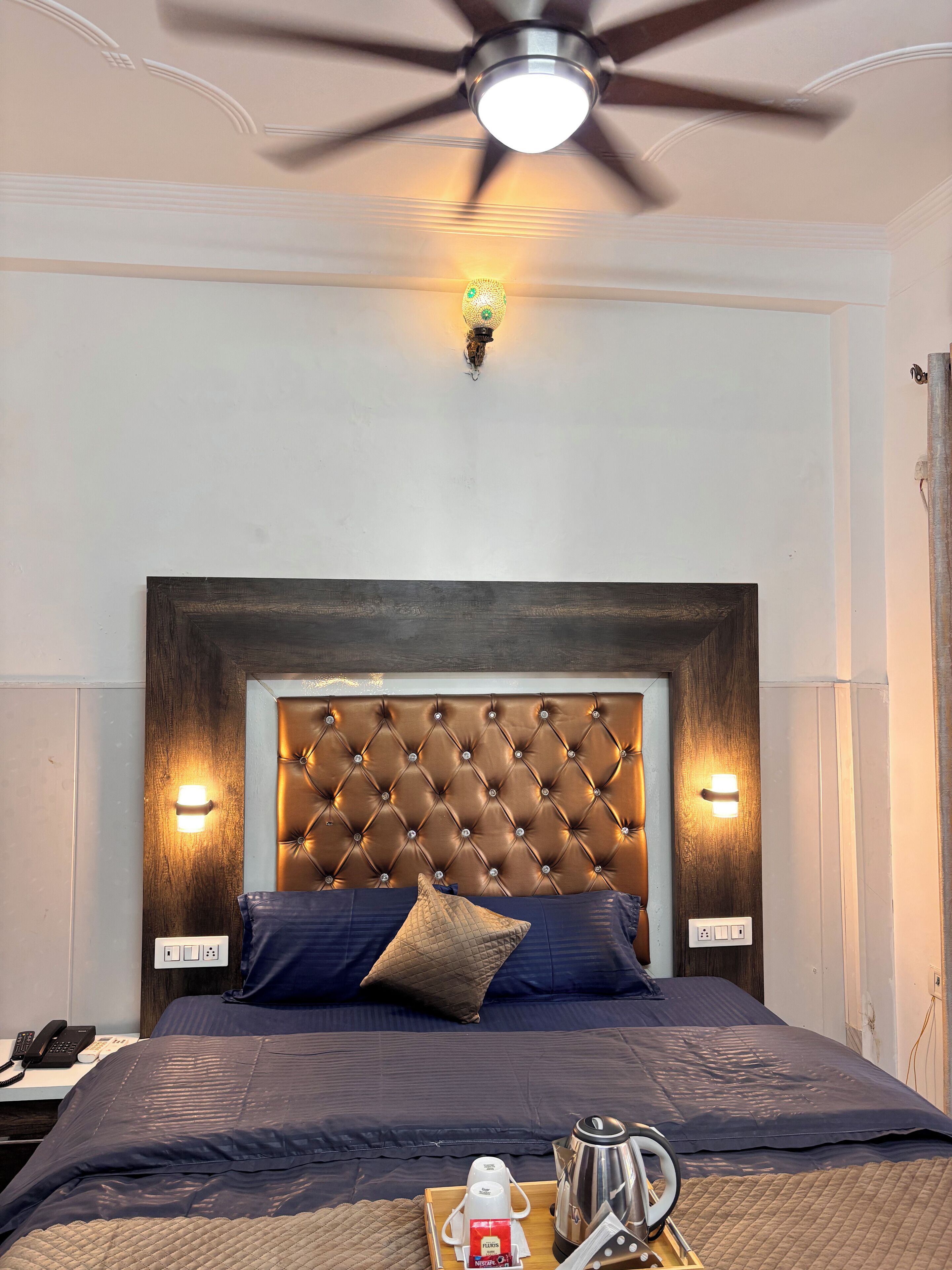 A hotel room with a tufted headboard, two wall-mounted reading lights, a ceiling fan, a bed with dark bedding, a bedside table with a phone, and a tray with coffee and tea.