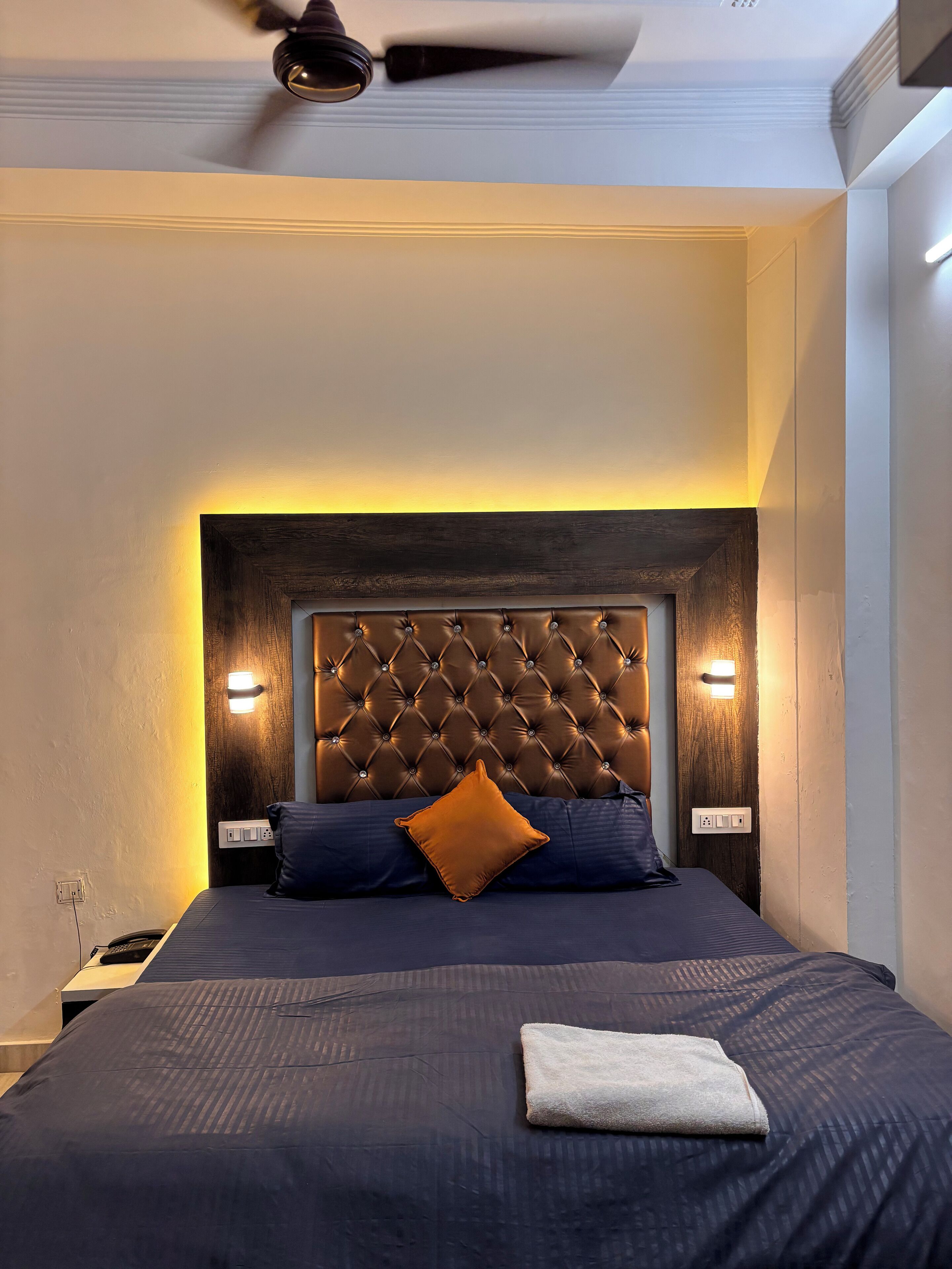 Un dormitorio con cabecera capitonada, dos lámparas de noche, una cama con ropa oscura y un ventilador de techo.