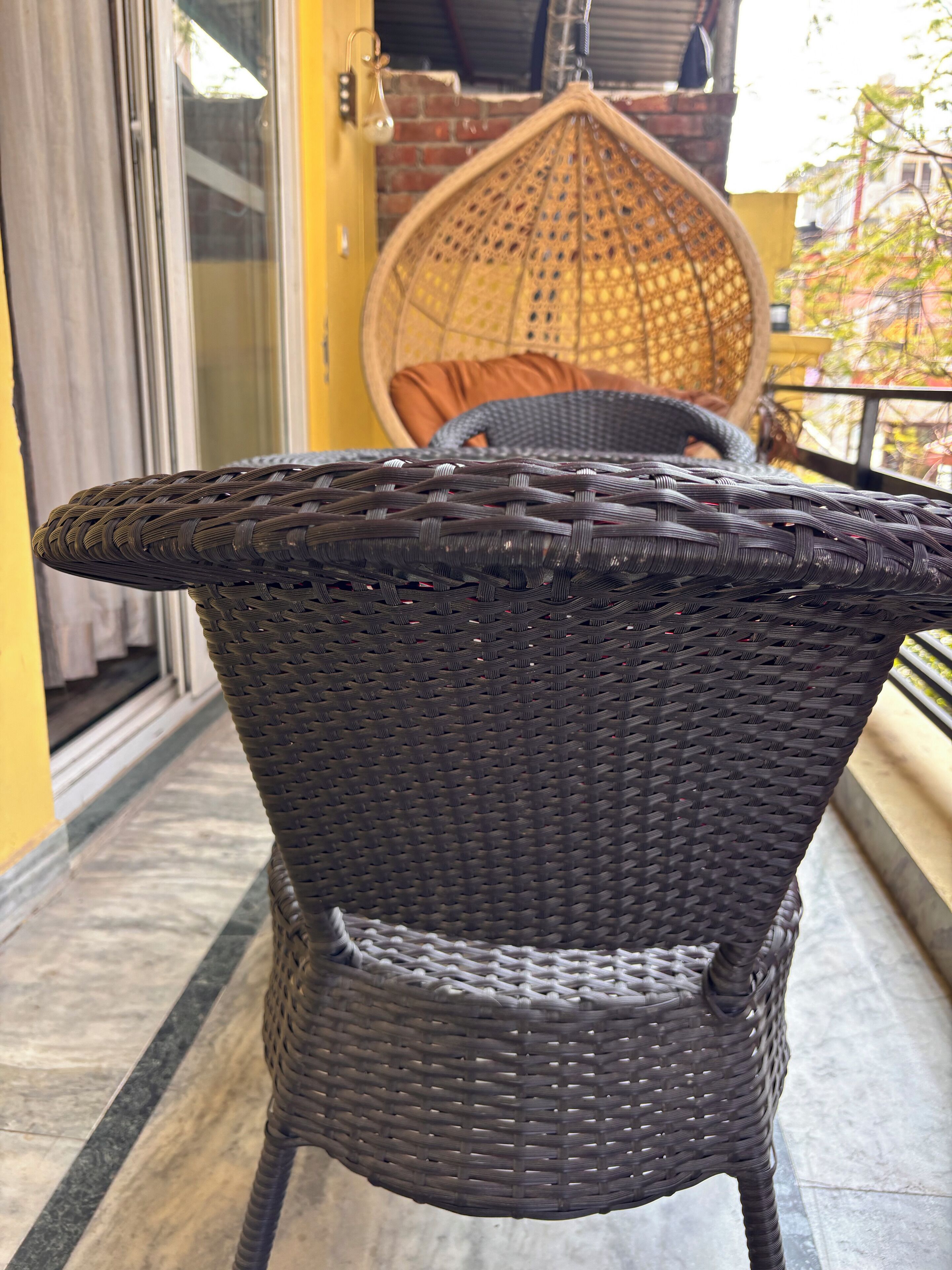 Une chaise en osier, avec une assise et un dossier tressés, placée sur un balcon d’où l’on aperçoit un immeuble et, au fond, un panier suspendu.