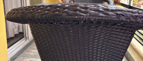 A wicker chair with a woven seat and backrest, positioned on a balcony with a view of a building and a hanging basket in the background.