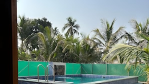 A swimming pool with tiled edges, surrounded by a green fence and palm trees. - SeaBreeze Nature Embrace Villa - Alibaug (Nagaon)