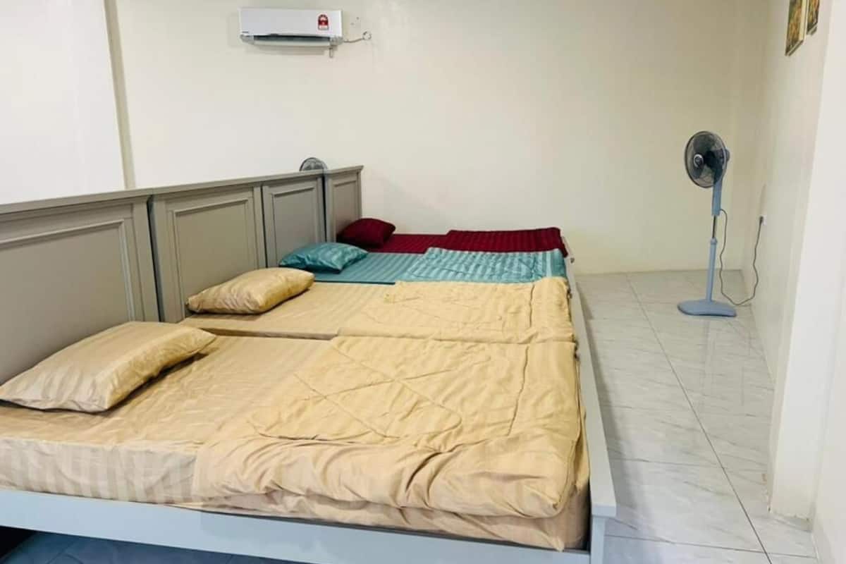 A bedroom with a bed, pillows, a bedside table, a wall-mounted air conditioner, and a standing fan.