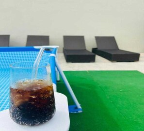 Un verre de cola posé sur une surface blanche, avec une table de billard bleue et des fauteuils noirs en arrière-plan.