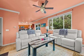 A spacious living room with a large window, a ceiling fan, and a kitchen in the background.