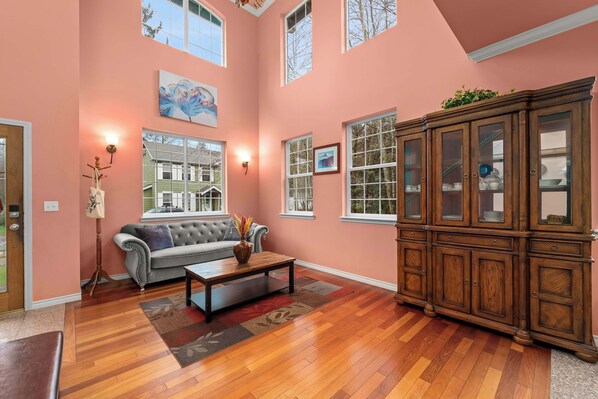 A spacious living room with hardwood flooring, a grey sofa, a wooden cabinet, and large windows.
