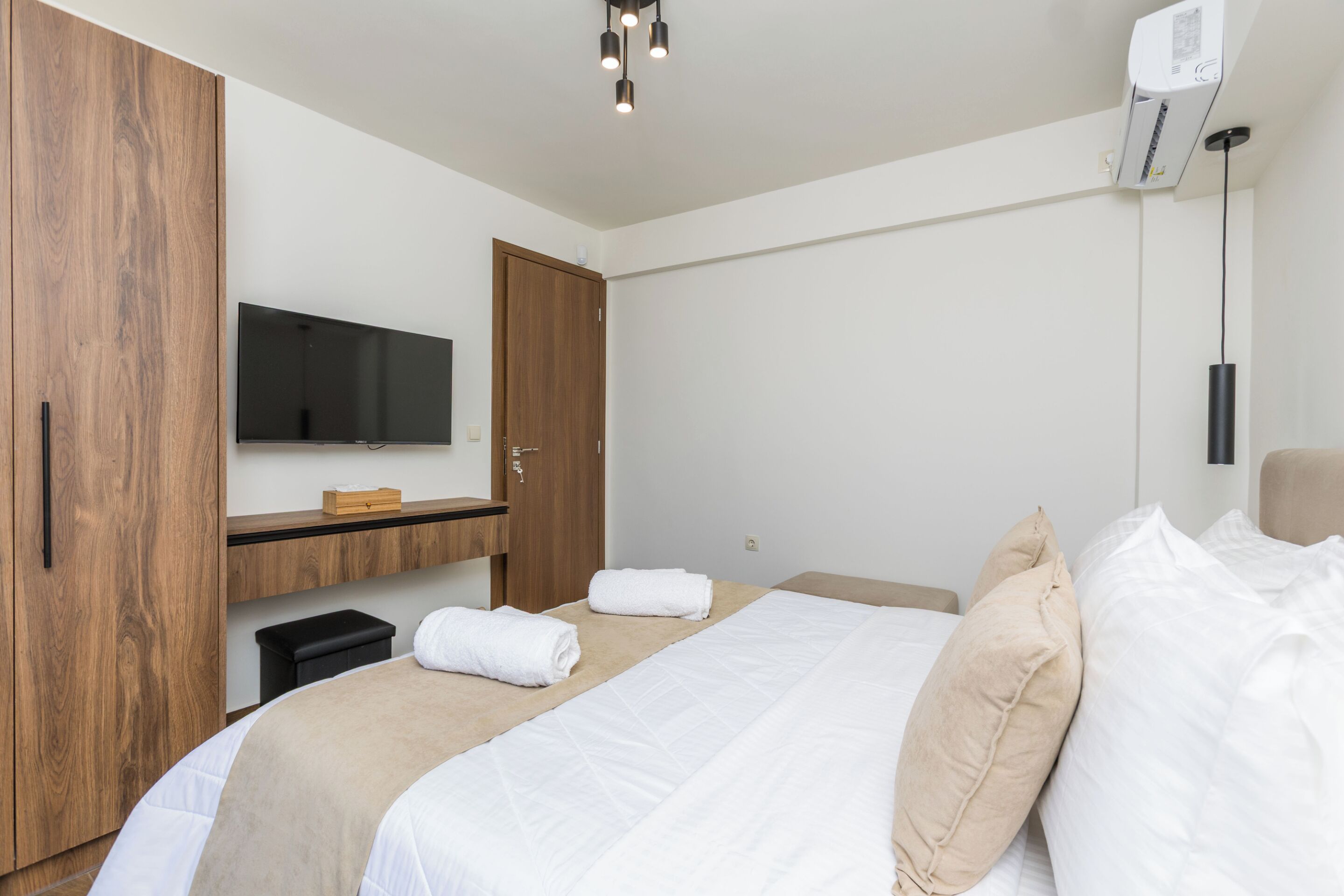 A modern hotel room with a large bed, a flat-screen TV mounted on the wall, a wooden wardrobe, and a small table with a box on it.