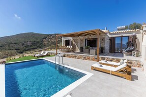 A modern outdoor pool area with lounge chairs and a pergola, surrounded by stone walls and a view of the hills. - Elithea Villas (Monemvasia)