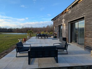 A modern patio with outdoor furniture, a large window, and a view of a lake. - Charming gîte in Sologne with private parking (Jouy-le-Potier)