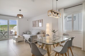 A modern living room with a glass dining table, grey chairs, a sofa, and a balcony with outdoor furniture. - Modern Atlanterra Apartment with Shared Pool, Solar Panels, Battery & Backup (Atlanterra)