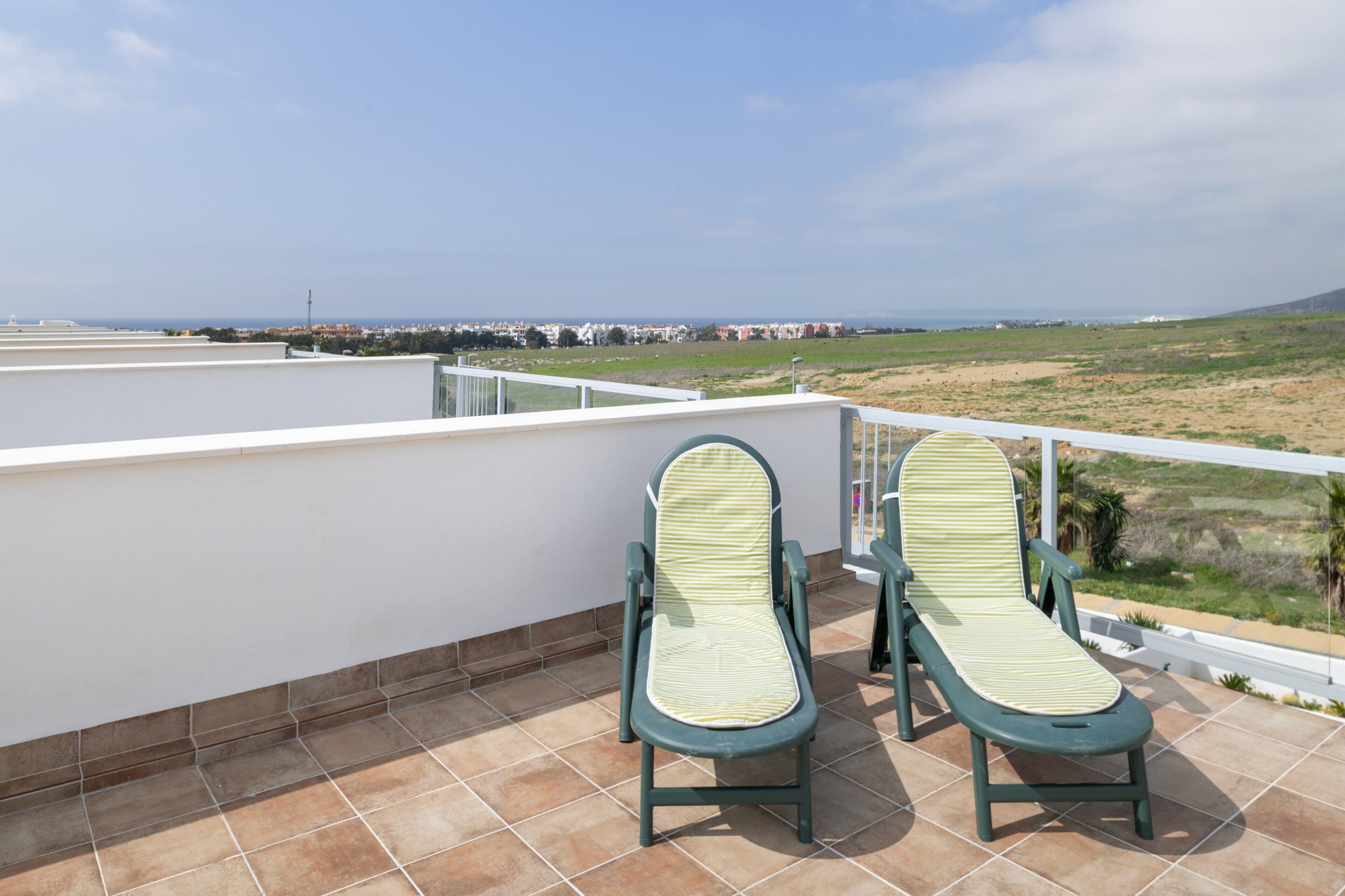 Une terrasse dotée de deux chaises longues, d’une balustrade blanche et offrant une vue sur la plage et les bâtiments situés au loin.