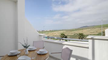 Un coin repas avec une table dressée pour quatre personnes, un vase agrémenté de verdure et une vue sur une plage et des collines.