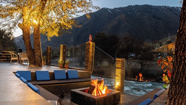 A fire pit with seating, a laptop, and a coffee maker on a terrace overlooking a river and mountains. - The Villa Himalaya (Srinagar)