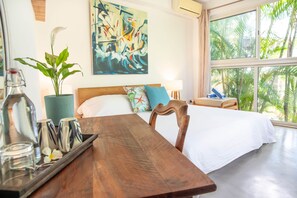 A hotel room with a bed, a wooden table, a potted plant, a painting on the wall, and a view of palm trees through the window. - 3 chambres privées avec panorama sur le Morne (Grande Riviere Noire)