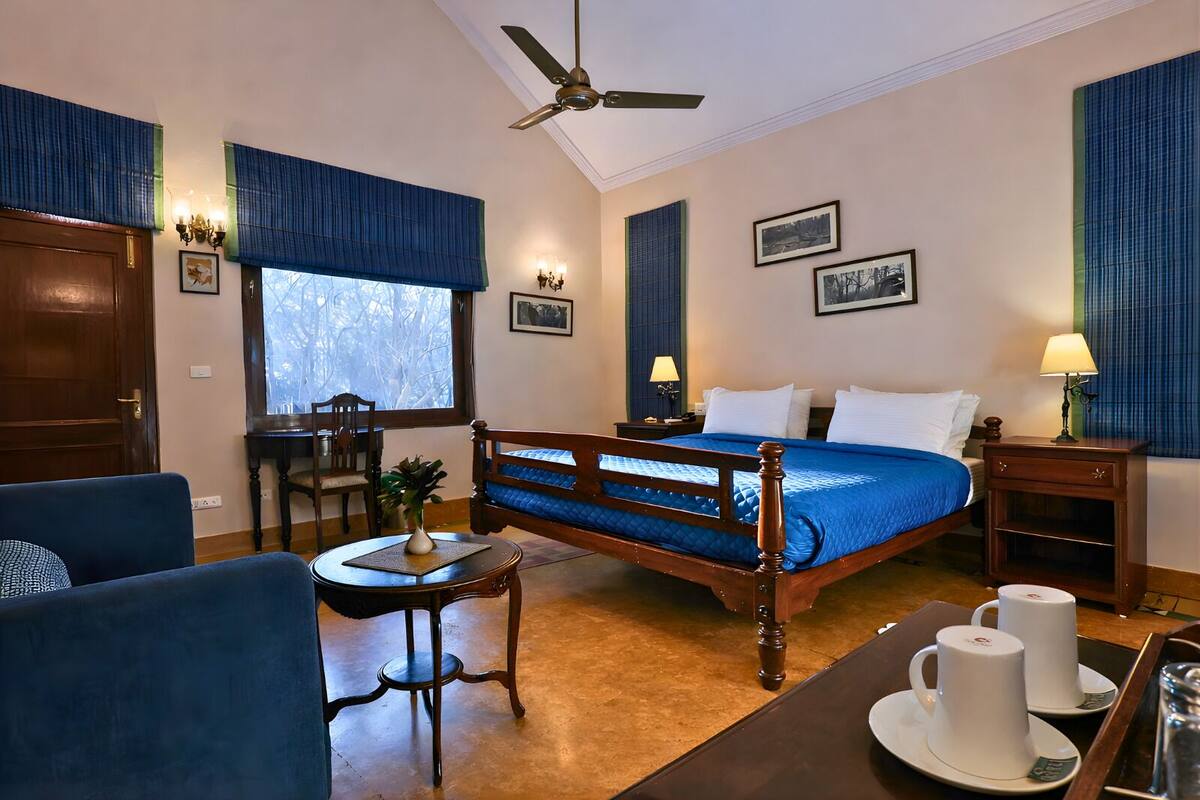 A hotel room with a bed, a desk, a chair, a small table with a vase, and a window with blinds.