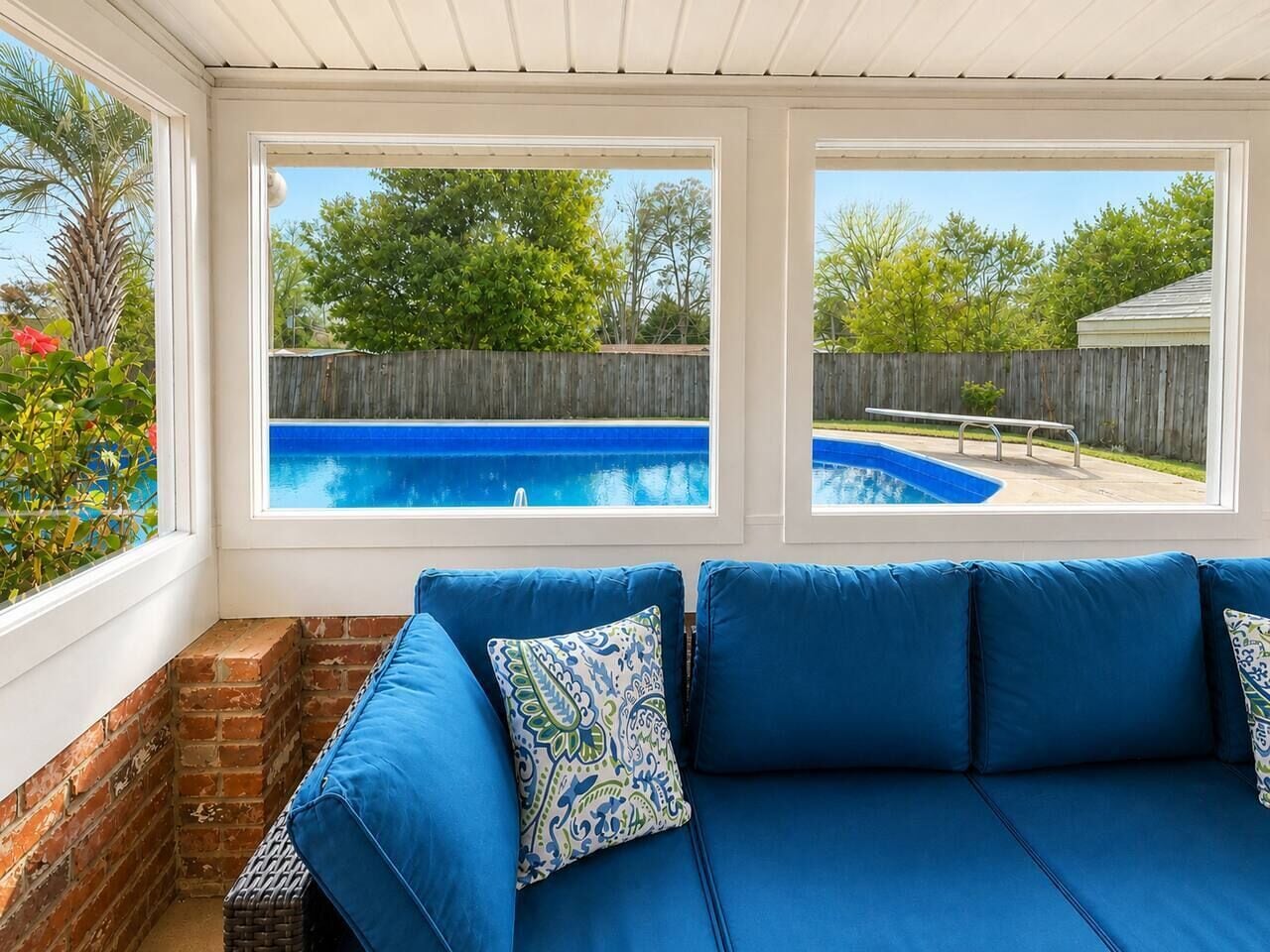 Una habitación con un sofá azul, dos ventanas con vistas a una piscina y una pared de ladrillo.