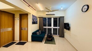 A living room with a teal sofa, a small table, a ceiling fan, and a clock on the wall. - 1BR Service Apart Parel 3504 Near TMH By Florastays (Mumbai)