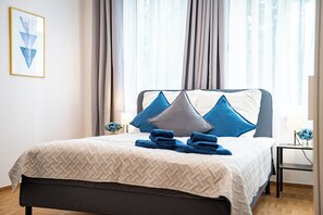 Un letto rifatto con cura, con una coperta a motivi geometrici, una testiera dal design geometrico e un comodino con una lampada e un vaso di fiori.