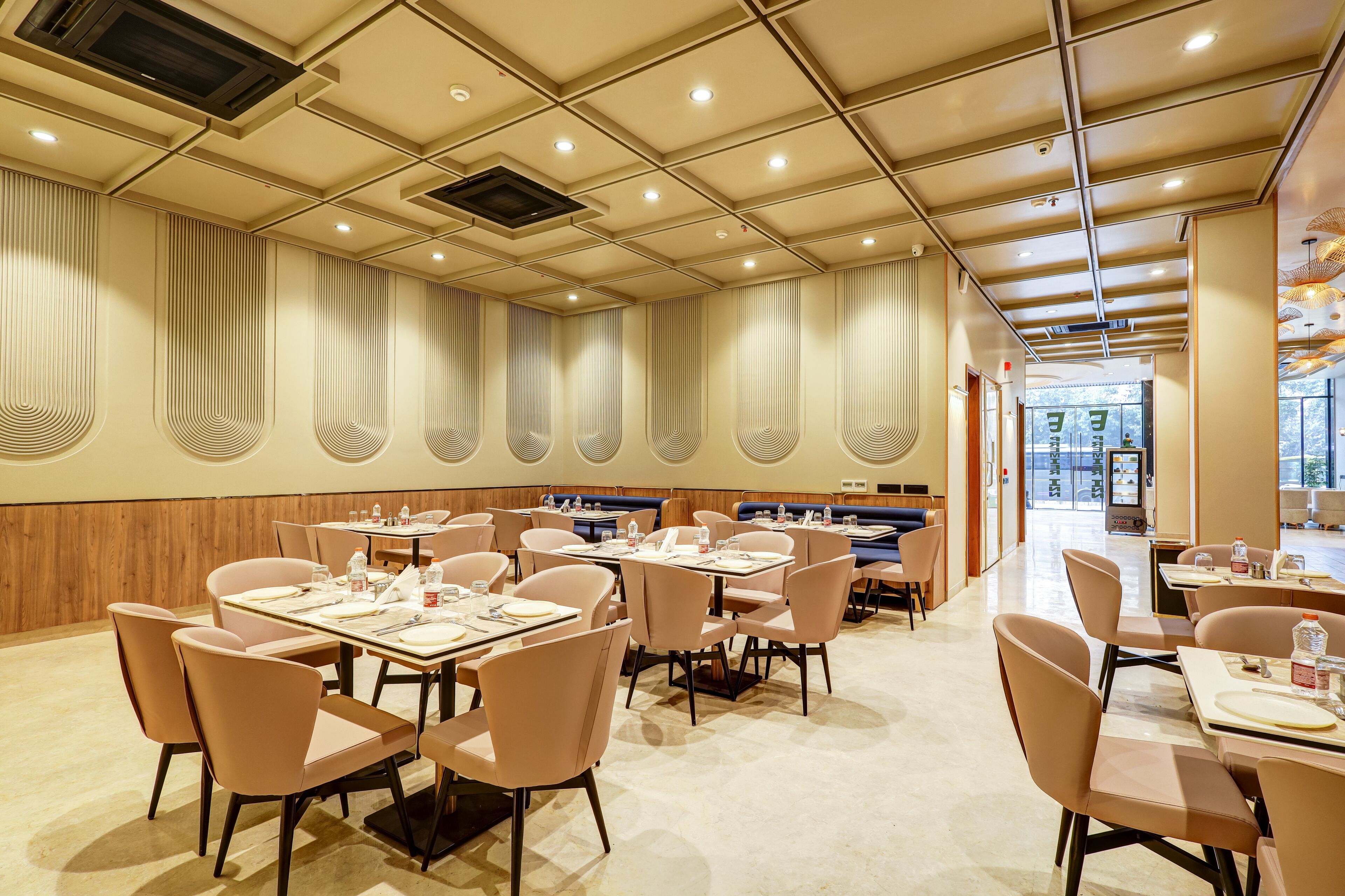 A spacious restaurant with high ceilings, recessed lighting, and a modern design. Tables are neatly arranged with chairs, and the walls feature decorative paneling.