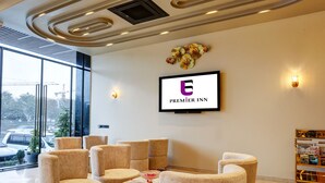 A modern lobby with a seating area, a television displaying the Premier Inn logo, and a view of the outside. - Hotel Premier Inn (New Delhi)