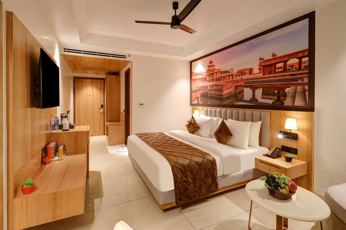 Habitación de hotel con cama, televisor, escritorio, mesita de noche y lámpara de pared.