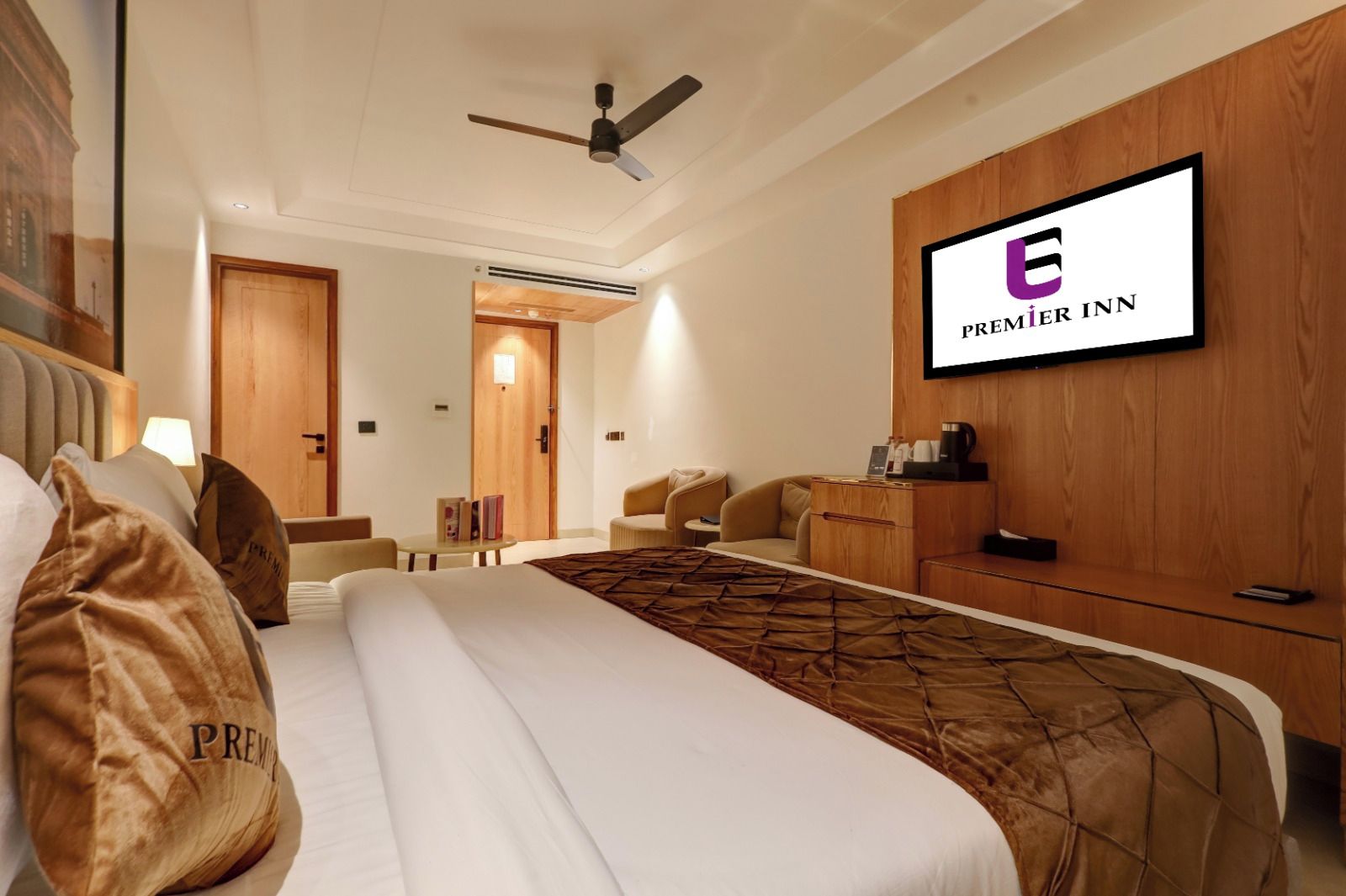 A hotel room with a bed, a television mounted on the wall, a ceiling fan, and a wooden door.