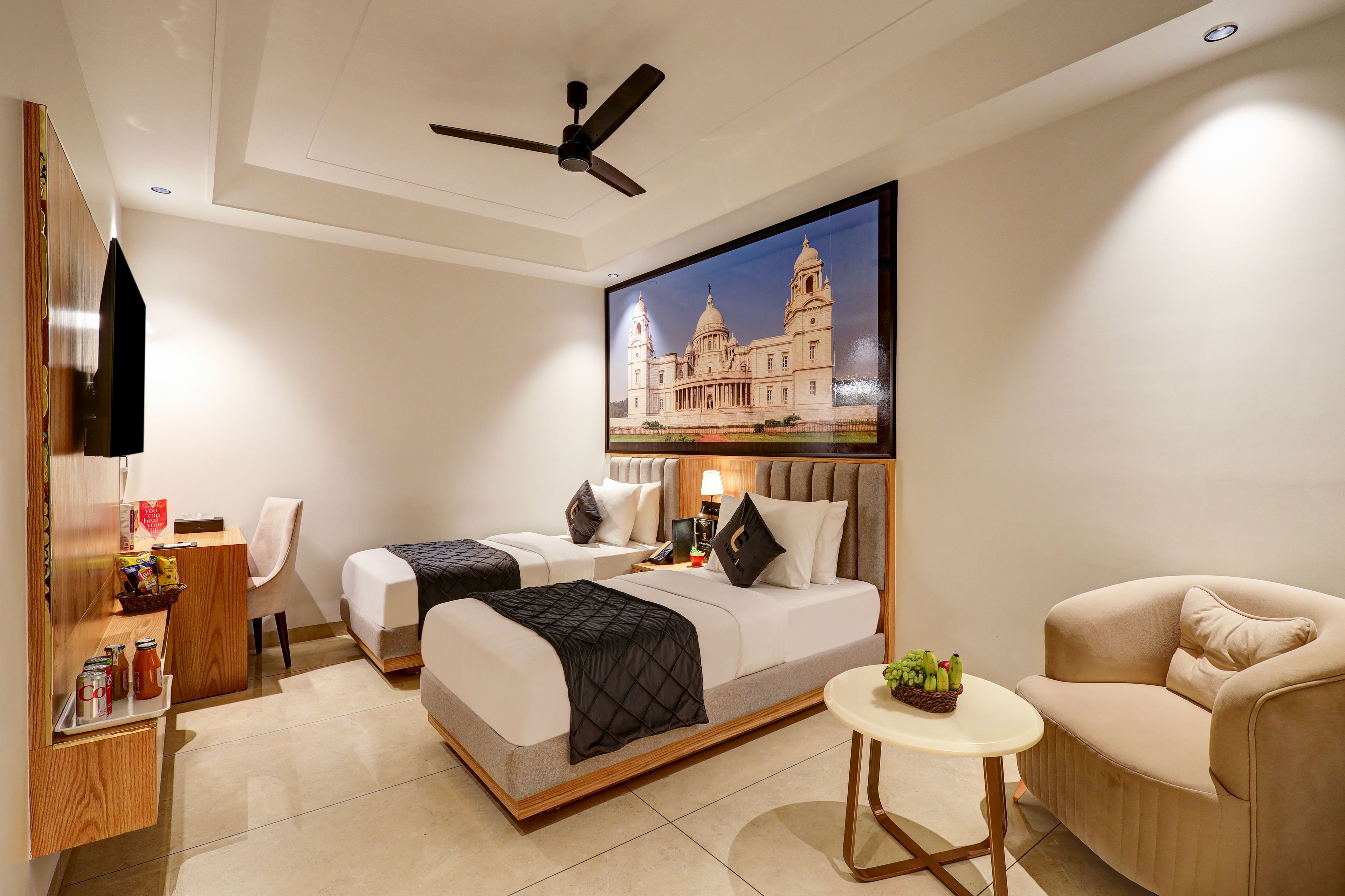 A hotel room with two beds, a desk, a chair, a small table with a plant, and a ceiling fan.