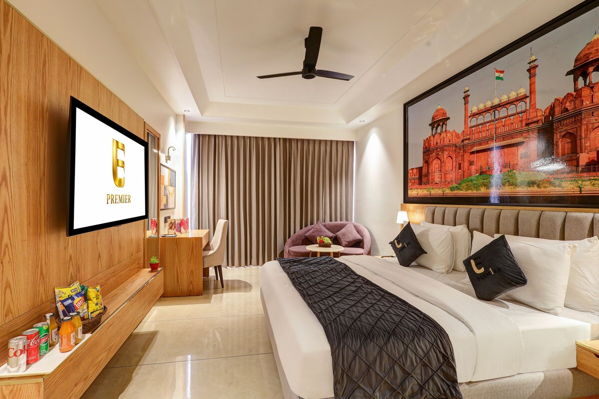 Una habitación de hotel con cama, televisor, ventilador de techo, un ventanal con cortinas y un cuadro colgado en la pared que muestra un edificio histórico.